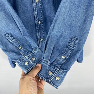 Op de afbeelding: Close-up van een blauw spijkeroverhemd. Het overhemd heeft een knoopsluiting, lange mouwen en manchetten met knopen. De knopen zijn zilverkleurig. De denimstof heeft een klassieke, medium-wash uitstraling.