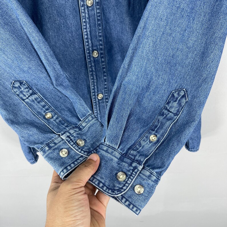 Op de afbeelding: Close-up van een blauw spijkeroverhemd. Het overhemd heeft een knoopsluiting, lange mouwen en manchetten met knopen. De knopen zijn zilverkleurig. De denimstof heeft een klassieke, medium-wash uitstraling.