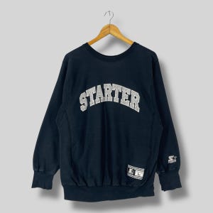 以下が含まれることがあります： 胸に白いアーチ状の文字で「STARTER」と書かれた黒いスターターのスウェットシャツ。クルーネック、長袖、袖に小さなスターターロゴが付いています。サイズ「L」のタグが見えます。