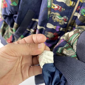 May include: A navy blue bomber jacket with a colorful pattern of horses and riders. The jacket has a zipper closure and a white tag with text that reads "U.S.A. 100% Nylon 377 7448".
