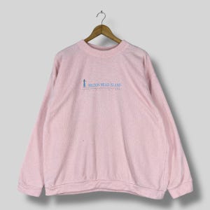 Può includere: Felpa rosa chiaro con la scritta "HILTON HEAD ISLAND" e un disegno di un faro blu. La felpa è appesa a una gruccia di legno.