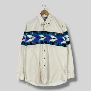May include: Cream-colored button-down shirt with a geometric pattern across the chest and sleeves. The pattern features blue, black, and white shapes. The shirt has long sleeves and a button-down collar.