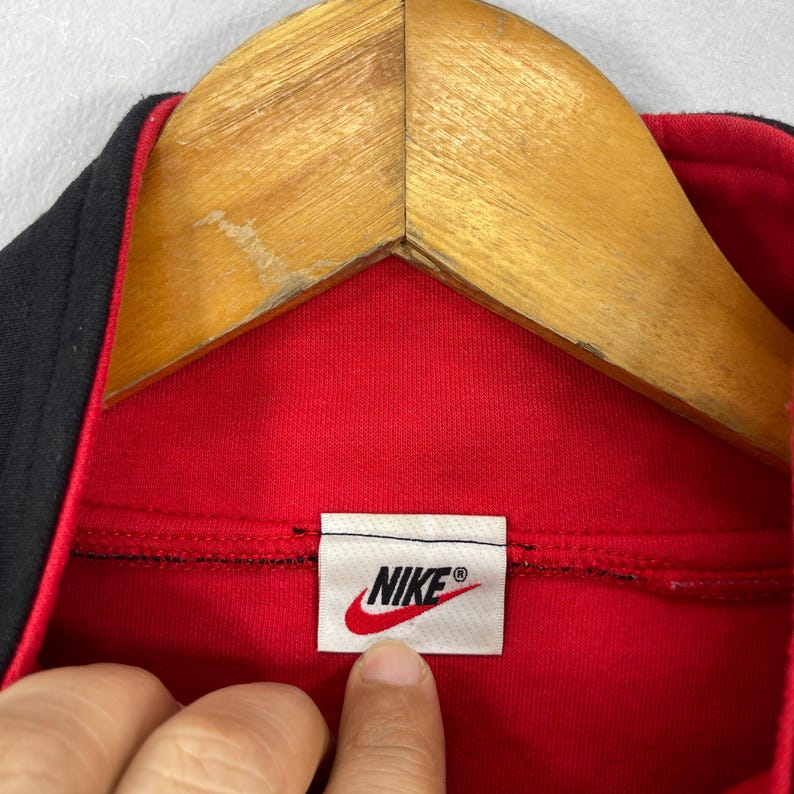 Może przedstawiać: Zbliżenie na czerwoną i czarną bluzę Nike z logo Nike w prawym dolnym rogu obrazu. Logo jest białe z czarnym znaczkiem.