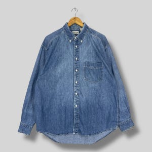 May include: A long-sleeved denim shirt in a medium blue wash, button-down style with a single chest pocket. The shirt is hanging on a wooden hanger. The collar has button-down points.