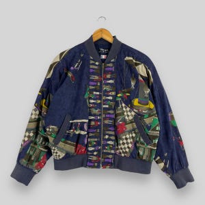 May include: A blue bomber jacket with a colorful, patterned design featuring a variety of figures and geometric shapes. The jacket has a zipper closure and a ribbed collar, cuffs, and hem.