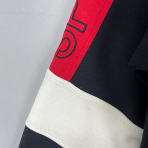 Może przedstawiać: Czarno-biało-czerwona bluza w paski z czarno-białym logo na czerwonym pasku.