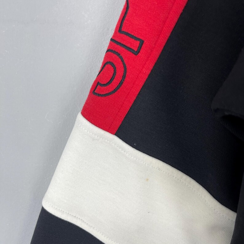 Może przedstawiać: Czarno-biało-czerwona bluza w paski z czarno-białym logo na czerwonym pasku.