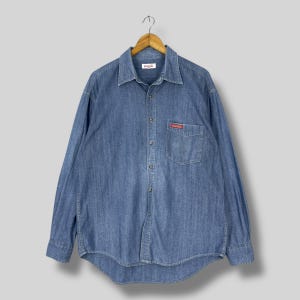 May include: A long-sleeved, light blue denim shirt with a button-down front and a collared neckline. The shirt features a small red Coca-Cola logo on the chest pocket and a Coca-Cola tag on the collar.