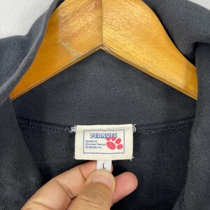 May include: Black sweatshirt with a tag featuring the Peanuts logo and the text "PEANUTS United Feature Syndicate, Inc." The tag also includes a paw print design. The sweatshirt is hanging on a wooden hanger.