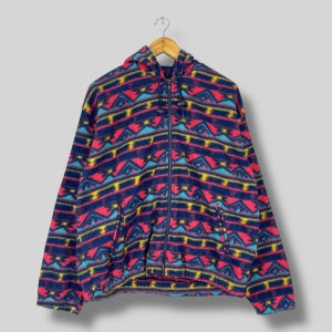 May include: A hooded fleece jacket with a full zipper, featuring a vibrant geometric pattern in navy blue, red, yellow, and turquoise. The jacket has two front pockets and is hanging on a wooden hanger.