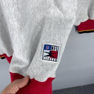 Puede incluir: Una sudadera gris claro con ribetes rojos y negros. La sudadera presenta un parche de NFL Fitness en la manga. Los pu&ntilde;os de las mangas son rojos con rayas negras y amarillas. La sudadera est&aacute; hecha de un material suave.
