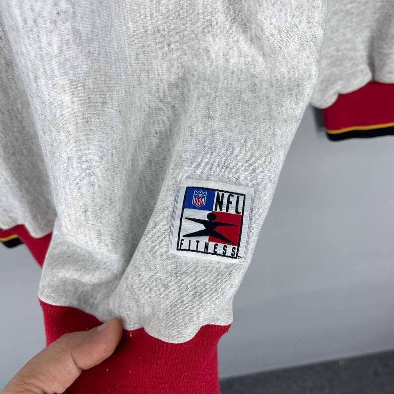 Puede incluir: Una sudadera gris claro con ribetes rojos y negros. La sudadera presenta un parche de NFL Fitness en la manga. Los pu&ntilde;os de las mangas son rojos con rayas negras y amarillas. La sudadera est&aacute; hecha de un material suave.