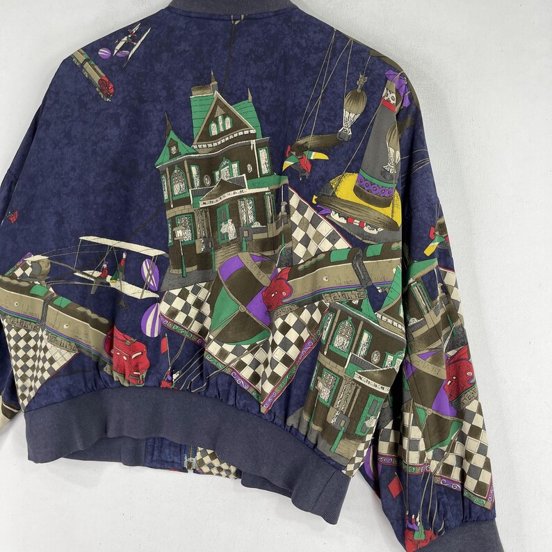 May include: A blue bomber jacket with a colorful, whimsical print featuring a house, a hot air balloon, and a checkered pattern.