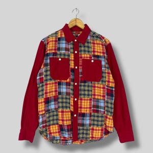 Puede incluir: Una camisa de manga larga con un diseño de patchwork. La camisa presenta un cuerpo y mangas rojas, con un patchwork de patrones de cuadros amarillos, azules y rojos en la parte delantera. Dos bolsillos rojos están en el pecho. La camisa está abotonada y colgada de una percha de madera.