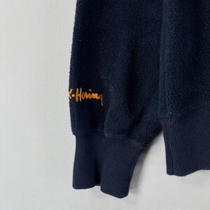 Puede incluir: Primer plano de una manga de sudadera de forro polar azul marino. El pu&ntilde;o y el cuerpo son de color azul marino liso. La firma del artista, "K-Haring", est&aacute; bordada con hilo naranja en la parte superior de la manga. La tela tiene un aspecto texturizado y difuso.