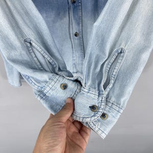 May include: Light blue denim shirt with buttoned cuffs and a button-down front. The shirt has a faded, ombre effect, transitioning from a darker blue at the top to a lighter shade towards the bottom. The cuffs are buttoned with small, round, brown buttons.