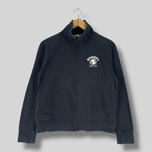 May include: A navy blue zip-up sweatshirt with a high collar. The sweatshirt features a white Snoopy logo with the text "SNOOPY SINCE 1950" on the chest. The jacket is hanging on a wooden hanger.