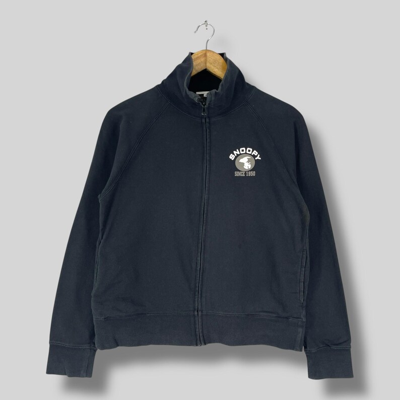 May include: A navy blue zip-up sweatshirt with a high collar. The sweatshirt features a white Snoopy logo with the text "SNOOPY SINCE 1950" on the chest. The jacket is hanging on a wooden hanger.