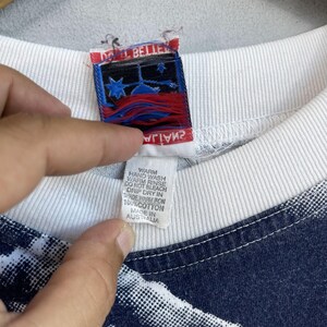 May include: A white t-shirt with a blue and white pattern. The tag says "Do It Belle" and has a red, white, and blue star design. The tag also says "Warm Hand Wash, Warm Rinse, Do Not Bleach, Drip Dry In Shade, Warm Iron, 100% Cotton, Made In Australia."