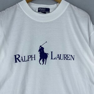 Puede incluir: Camiseta blanca con un logotipo de Polo Ralph Lauren azul marino que representa a un jugador de polo a caballo. El logotipo tambi&eacute;n incluye el texto "Ralph Lauren" en azul marino.