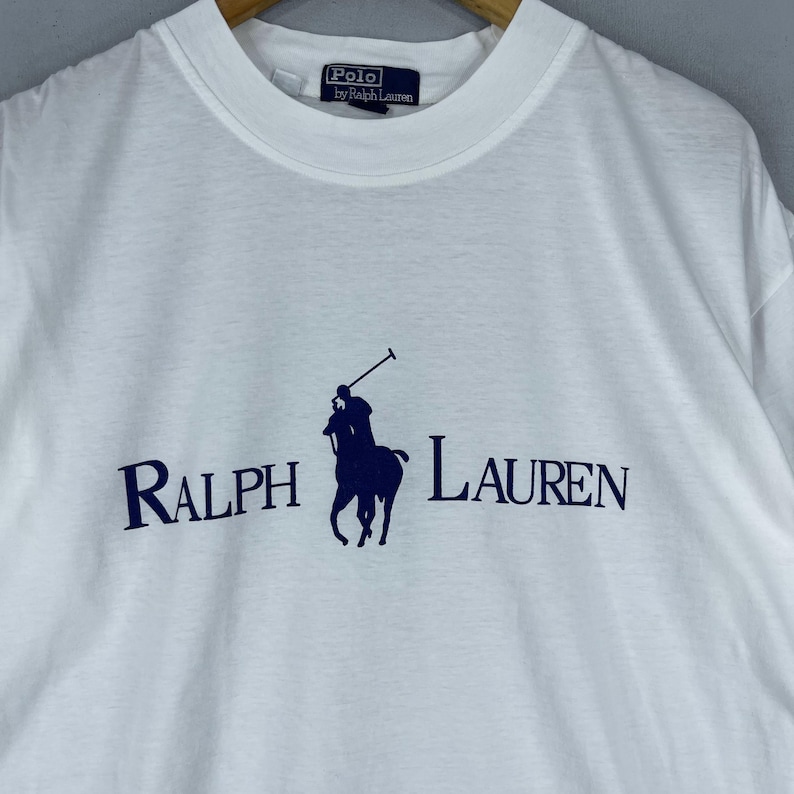 Puede incluir: Camiseta blanca con un logotipo de Polo Ralph Lauren azul marino que representa a un jugador de polo a caballo. El logotipo tambi&eacute;n incluye el texto "Ralph Lauren" en azul marino.