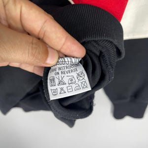 Może przedstawiać: Biała metka z czarnym tekstem, kt&oacute;ry brzmi "Care Instructions on Reverse" i r&oacute;żnymi symbolami dotyczącymi instrukcji prania. Metka jest przymocowana do czarnej bluzy.