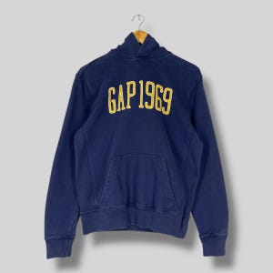 Puede incluir: Sudadera con capucha azul marino con el texto "GAP 1969" en amarillo en el pecho. La sudadera tiene un bolsillo de canguro y está colgada de una percha metálica. La prenda está hecha de un material suave.