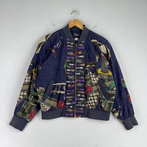 May include: A navy blue bomber jacket with a colorful patterned design. The jacket has a zipper closure and two pockets. The pattern features a variety of geometric shapes and figures, including a checkered pattern, stripes, and a whimsical design of people and objects.