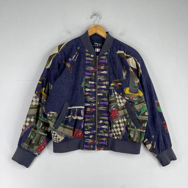 May include: A navy blue bomber jacket with a colorful patterned design. The jacket has a zipper closure and two pockets. The pattern features a variety of geometric shapes and figures, including a checkered pattern, stripes, and a whimsical design of people and objects.