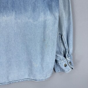 May include: Light blue denim shirt with long sleeves. The shirt features a button-down front and cuffs, with a faded wash effect. The fabric has a textured appearance, and the shirt is in good condition.