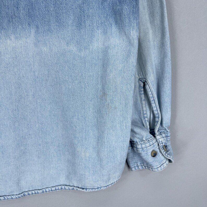 May include: Light blue denim shirt with long sleeves. The shirt features a button-down front and cuffs, with a faded wash effect. The fabric has a textured appearance, and the shirt is in good condition.