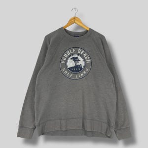 May include: Gray sweatshirt with a crew neck and long sleeves. The front features a circular graphic with the text "PEBBLE BEACH GOLF LINKS 1919" in navy blue and white. The sweatshirt is hanging on a wooden hanger.