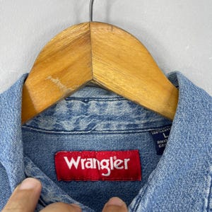 May include: Close-up of a light blue denim Wrangler shirt. The red Wrangler label is prominently displayed. The shirt is hanging on a wooden hanger. The shirt is made of 100% cotton.
