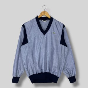 May include: A light blue and navy blue long-sleeve pullover shirt with a V-neck. The shirt has navy blue accents on the shoulders, cuffs, and hem. A small green crocodile logo is on the chest.