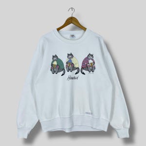 Op de afbeelding: Witte sweatshirt met een afbeelding van drie cartoonkatten die elk een ijsje vasthouden. De katten zijn grijs met kleurrijke shirts. Het woord "Hawaii" staat eronder in cursief. De sweatshirt hangt aan een houten hanger.