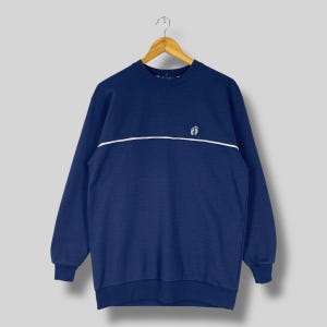 Puede incluir: Una sudadera azul marino con una raya horizontal blanca en el pecho. La parte superior de manga larga tiene cuello redondo y un pequeño logotipo encima de la raya. La sudadera está colgada en una percha de madera.