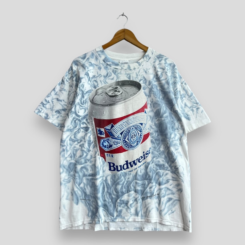 May include: A white t-shirt with a tie-dye pattern featuring a large image of a Budweiser beer can. The can is silver with a red, white, and blue label. The text "Budweiser" is printed on the label.
