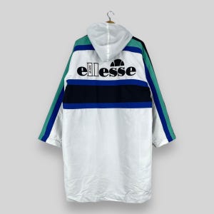 Op de afbeelding: Witte capuchontrui met een blauw, zwart en groen gestreept patroon. De trui heeft het woord "ellesse" in zwarte letters op de rug.