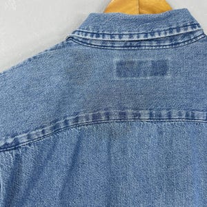 May include: Close-up of the back of a light blue denim shirt. The collar and yoke are visible, with a rectangular patch near the yoke. The denim fabric has a textured appearance.