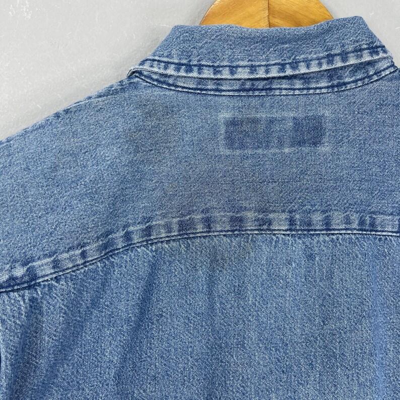 May include: Close-up of the back of a light blue denim shirt. The collar and yoke are visible, with a rectangular patch near the yoke. The denim fabric has a textured appearance.
