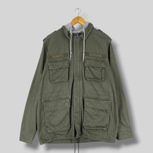 May include: Olive green hooded jacket with multiple front pockets and the text "USANYC" and "DIVISION". The jacket has a gray hood and white drawstrings. It is hanging on a wooden hanger.