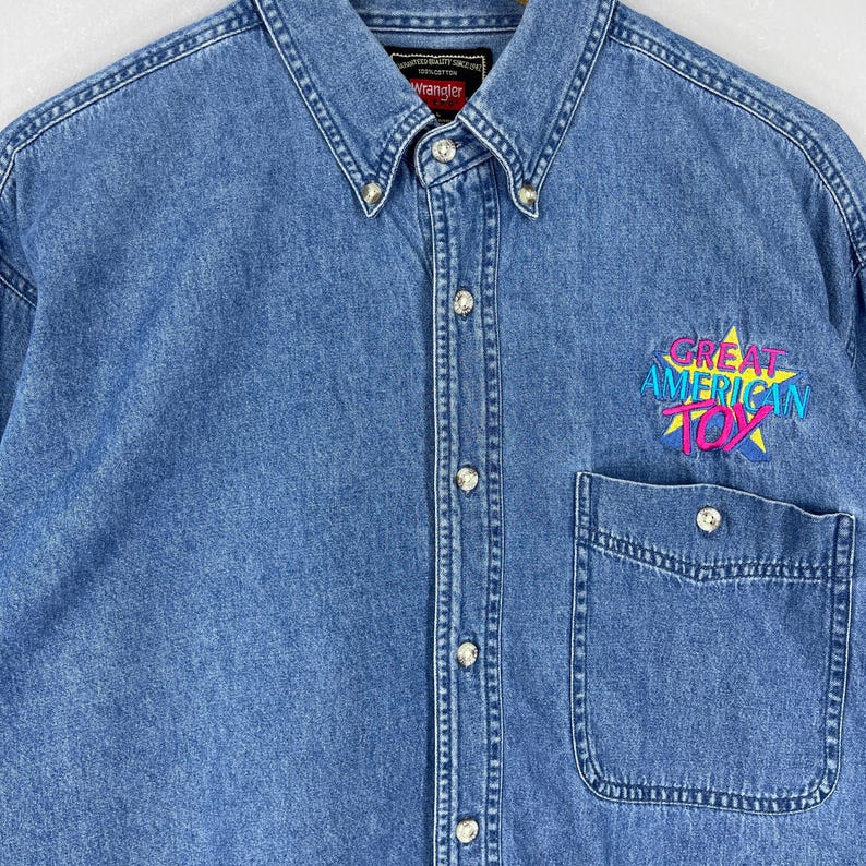 Op de afbeelding: Lichtblauwe denim overhemd met knoopsluiting en borstzak. De tekst "GREAT AMERICAN TOY" staat in een sterontwerp op het overhemd. Het Wrangler-merklabel is zichtbaar op de kraag.