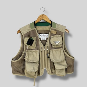 Op de afbeelding: Tan Columbia visvest met meerdere zakken, mesh zijkanten en een ritssluiting. Het vest heeft een groene kraag en een klein zwart accessoire bevestigd aan een zak. Het Columbia-logo is zichtbaar.