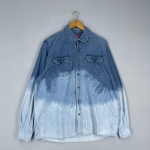 May include: A long-sleeved denim shirt with a two-tone blue wash. The shirt features two chest pockets, a button-down front, and a classic collar. The bottom half of the shirt fades to a lighter blue.