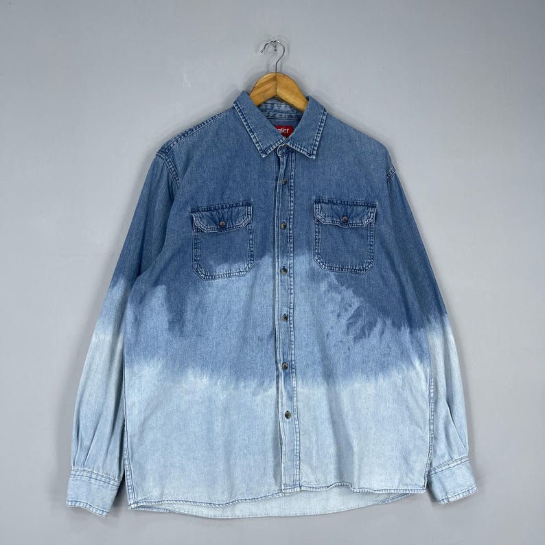 May include: A long-sleeved denim shirt with a two-tone blue wash. The shirt features two chest pockets, a button-down front, and a classic collar. The bottom half of the shirt fades to a lighter blue.