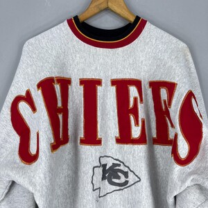 Puede incluir: Sudadera gris con la palabra "CHIEFS" en letras rojas en el pecho. Las letras tienen un contorno dorado. Un logotipo del equipo est&aacute; debajo del texto. El cuello es negro con ribete dorado y rojo.