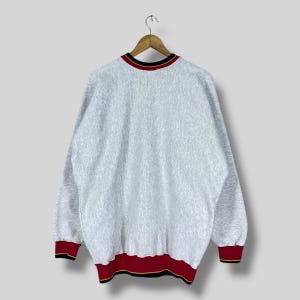 Puede incluir: Una sudadera gris jaspeada con cuello redondo y mangas largas. El cuello, los pu&ntilde;os y el dobladillo presentan un dise&ntilde;o a rayas rojas, negras y doradas. La sudadera est&aacute; colgada de una percha de madera.