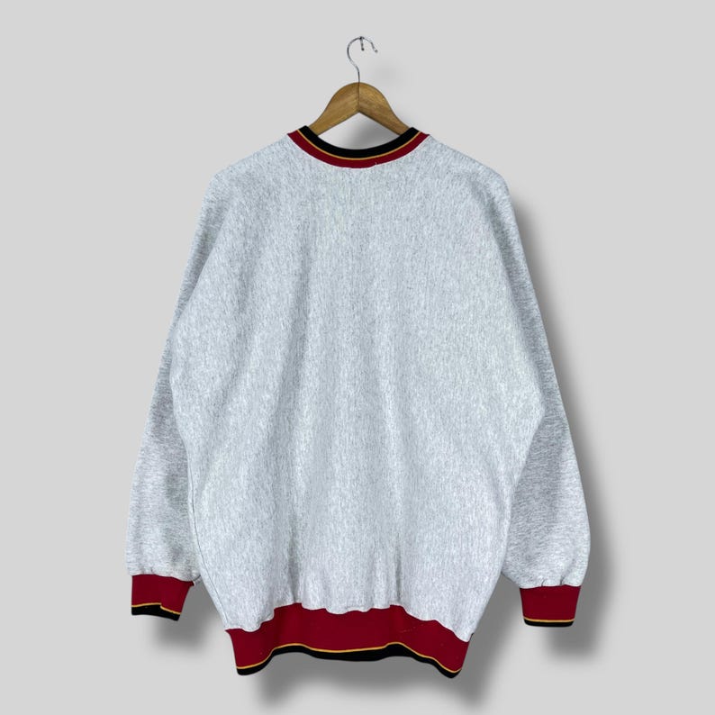 Puede incluir: Una sudadera gris jaspeada con cuello redondo y mangas largas. El cuello, los pu&ntilde;os y el dobladillo presentan un dise&ntilde;o a rayas rojas, negras y doradas. La sudadera est&aacute; colgada de una percha de madera.