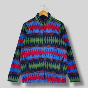 May include: A full-zip fleece jacket with a vibrant geometric pattern in red, blue, green, and navy. The jacket has a stand-up collar, two side pockets, and a soft, textured appearance. The jacket is hanging on a wooden hanger.