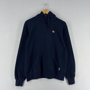 Puede incluir: Sudadera con capucha azul marino con cord&oacute;n y bolsillo tipo canguro. La sudadera tiene un peque&ntilde;o logotipo bordado en el pecho y un logotipo en el pu&ntilde;o. La sudadera con capucha est&aacute; colgada de una percha de madera.